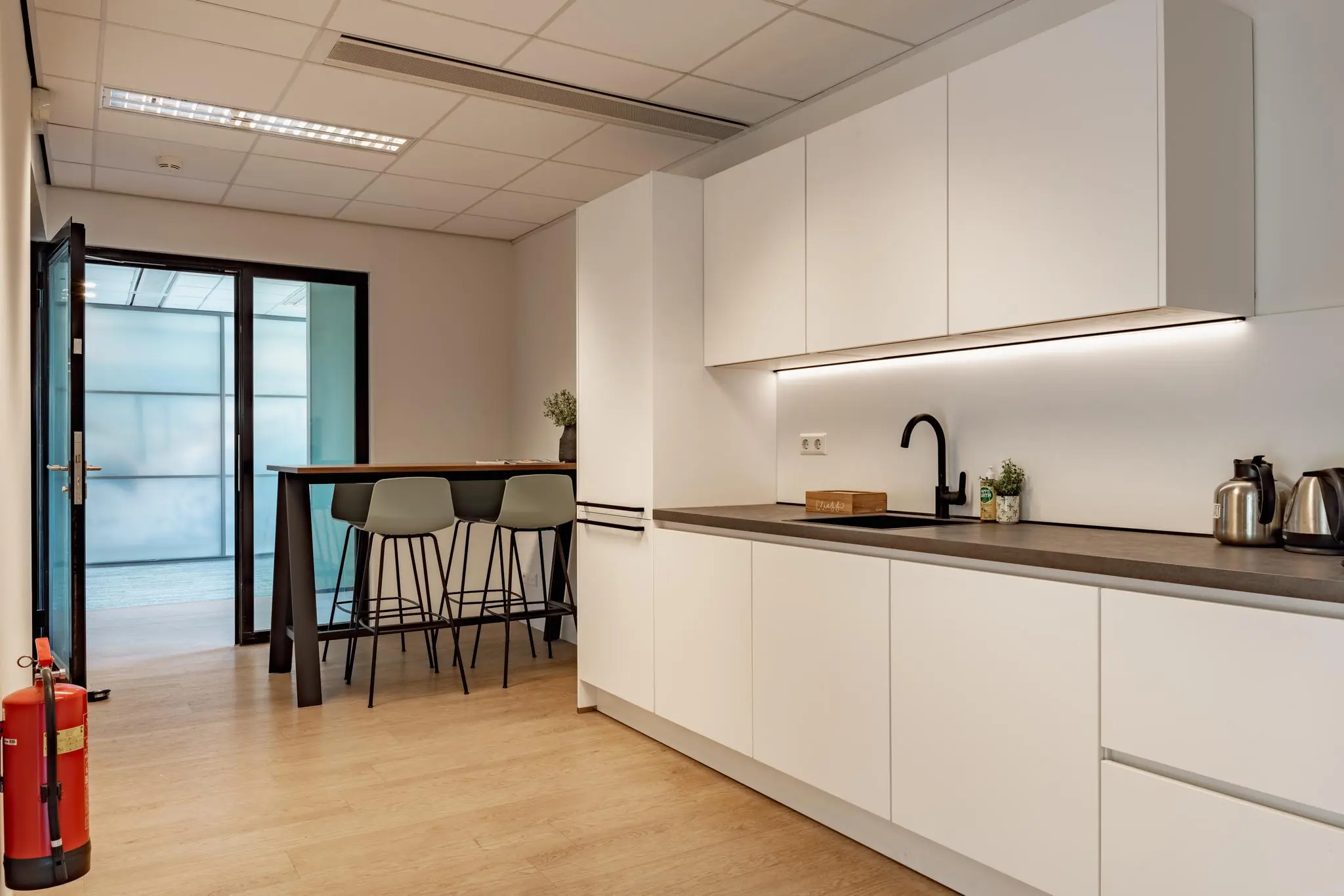 Moderne kantoorpantry met witte kasten, een zwarte kraan, koffiezetapparaten en een hoge tafel met barkrukken.